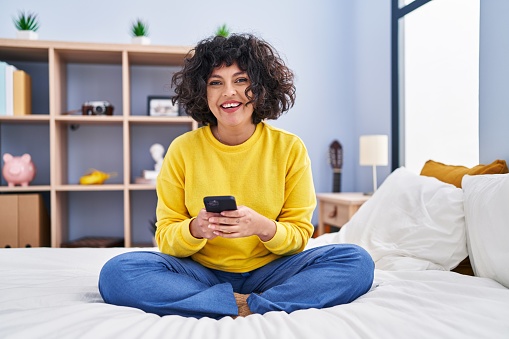 Young beautiful hispanic woman using smartphone sitting on bed at bedroom