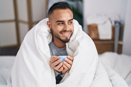 Young hispanic man drinking cup of coffee sitting on bed at bedroom