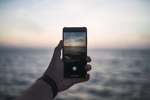 A closeup of a male's hand taking a picture of a beautiful sunset with his phone
