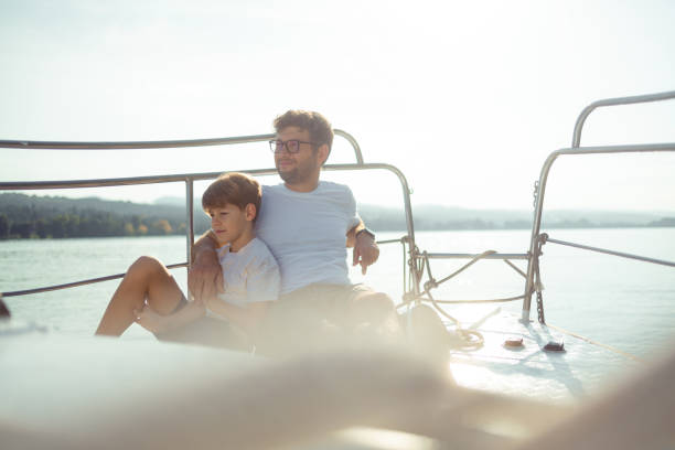 Father and son enjoying a day on the water. They sit on the bow of the ship and look into the distance stock photo