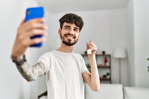 Young hispanic man making selfie by the smartphone holding key of new home