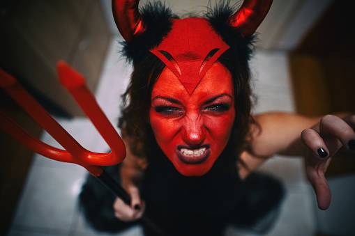Woman dressed in a devil's outfit for Halloween looking at the camera while making a claw with one hand and holding trident in another.