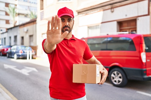 Young hispanic man with beard wearing delivery uniform and cap holding box with open hand doing stop sign with serious and confident expression, defense gesture