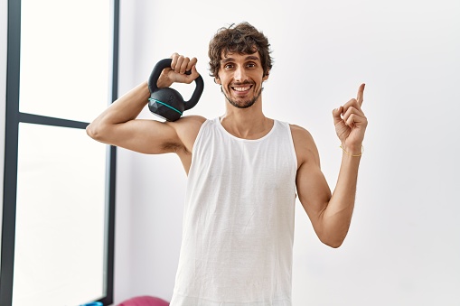 Young hispanic man wearing sportswear using dumbbells smiling happy pointing with hand and finger to the side