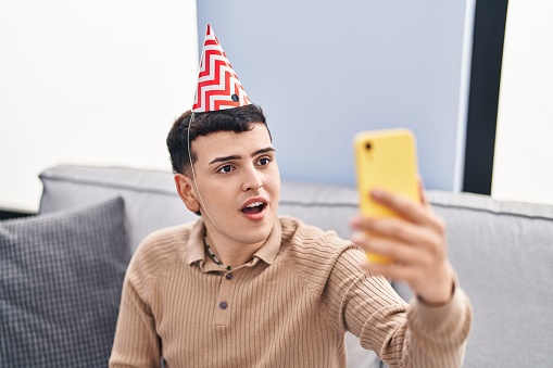 Non binary person celebrating birthday doing video call scared and amazed with open mouth for surprise, disbelief face