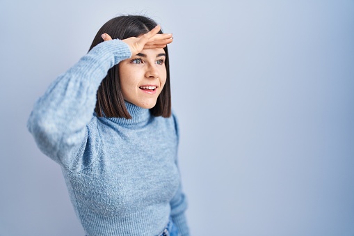 Young hispanic woman standing over blue background very happy and smiling looking far away with hand over head. searching concept.