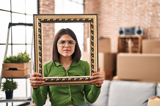 Young hispanic woman at new home holding empty frame skeptic and nervous, frowning upset because of problem. negative person.