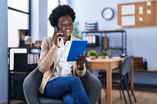 Young african american woman business worker using touchpad talking on the smartphone at office