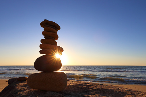 Several small stones balance on a large boulder at a beach as the sun is about to set behind the small tower of rocks.