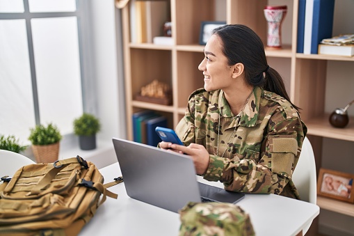 Young hispanic woman army soldier using laptop and smartphone at home