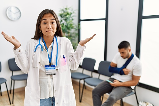 Young asian doctor woman at waiting room with a man with a broken arm clueless and confused expression with arms and hands raised. doubt concept.