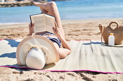 Young hispanic woman wearing bikini and summer hat reading book at seaside