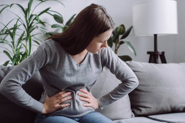Sick young woman in pain holding belly stomach feeling hurt abdomen ache gastritis pancreatitis symptoms. Upset girl sitting on couch suffering from indigestion flatulence abdominal problem concept stock photo