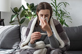 Seasonal contagious grippe illness concept. Ill woman covered plaid feel sick unwell. Unhealthy female measuring body temperature with electronic thermometer, suffer from flu sitting on sofa at home