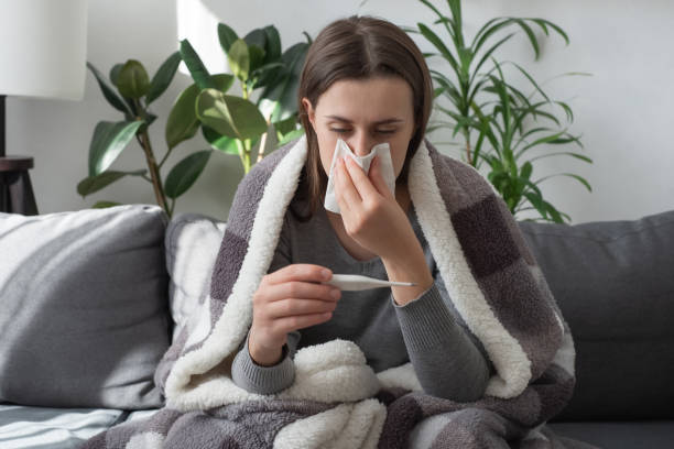 Seasonal contagious grippe illness concept. Ill woman covered plaid feel sick unwell. Unhealthy female measuring body temperature with electronic thermometer, suffer from flu sitting on sofa at home stock photo