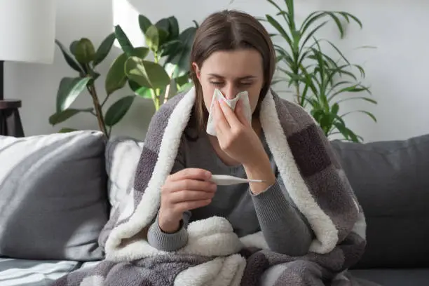 Photo of Seasonal contagious grippe illness concept. Ill woman covered plaid feel sick unwell. Unhealthy female measuring body temperature with electronic thermometer, suffer from flu sitting on sofa at home