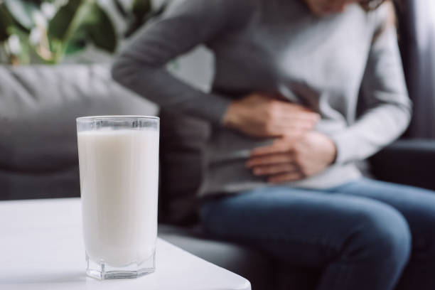 Lactose intolerance, food allergy concept. Selective focus of glass milk is on white table and unhealthy young female touch stomach suffers from severe ache sitting on couch in living room at home Lactose intolerance, food allergy concept. Selective focus of glass milk is on white table and unhealthy young female touch stomach suffers from severe ache sitting on couch in living room at home lactose fermentation stock pictures, royalty-free photos & images
