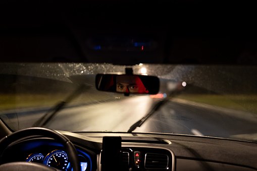 Eyes of young man are reflected in rearview mirror during night trip around city. Taxi driver or evening courier is driving along dark highway in passenger car with blue backlight of speedometer