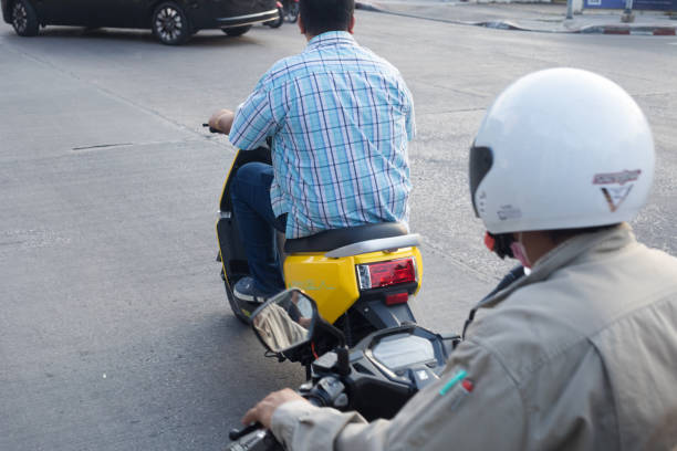 Thai man is driving on electric motorcycle. Scene is at junction in Bangkok Ladprao. Man is is driving over junction and is sitting on new yellow eletric motorcycle. He is wearinf casual clothing and no crash helmet. Another person with crash helmet is following