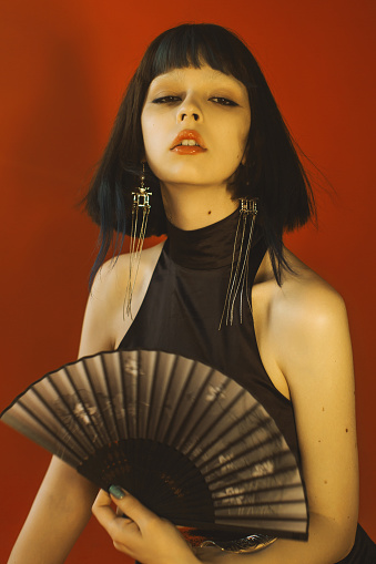 Beauty portrait. Hairstyle. Brunette asian woman with hand fan posing in studio on red background. Fashion model with pale skin and brown hair in black dress. Style. Earrings