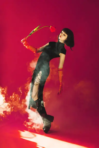 Photo of Brunette asian woman with tulip in hand posing with smoke in studio on red background. Fashion model with pale skin and brown hair in black dress. Gloves on hands. Style