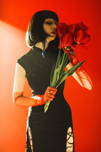 Brunette asian woman with tulip in hand posing with smoke in studio on red background. Fashion model with pale skin and brown hair in black dress. Gloves on hands. Style