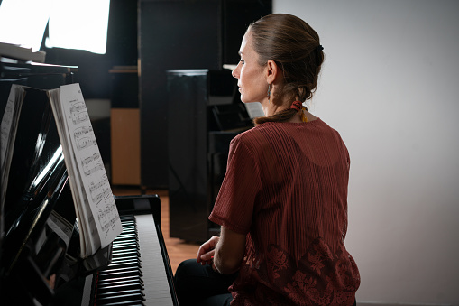 Back view of adult caucasian woman preparing to play piano at a concert. It is placed on a theater stage and plays a grand concert grand piano. Partially backlit, . Black spotlights in the background.