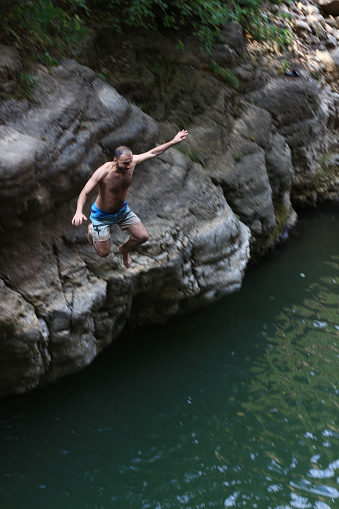 A vertical shot of an excited screaming Caucasian man in swimsuit jumping into a forest lake from a rough rugged cliff