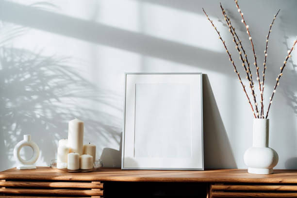 istock Modern minimalist style interior with white poster mockup, candles and blooming branches of the pussy willow in vase on wooden console under sunlight and shadows on a white gray wall. Selective focus. 1437974169