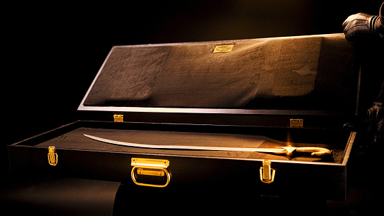 Elegant antique Venetian sword in black leather case. Close up of male hands in black gloves opening a case with a valuable vintage sabre on black background under the dim light.