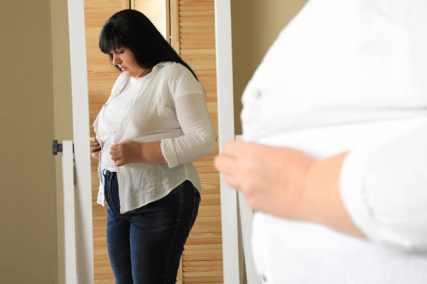 太りすぎの女性が自宅の鏡の前でタイトなシャツのボタンを留めようとしている - mature adult sadness overweight women ストックフォトと画像