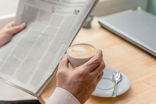 close-up of the hands of an unrecognizable older man holding a newspaper in one hand and a cup of coffee in the other, a closed laptop on the table, concept of leisure