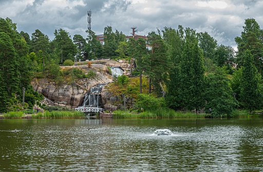 Finland, Kotka - July 18, 2022: Sopokanlahti park and lake. the waterfall from across the lake set in green environment under gray cloudscape. Building and antenna on horizon