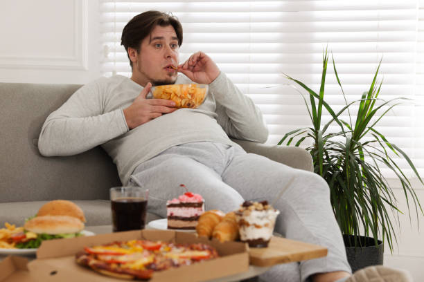 Overweight man with chips on sofa at home Overweight man with chips on sofa at home over eating stock pictures, royalty-free photos & images