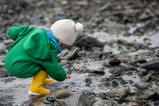 Cute little girl wearing green jumpsuit and yellow rubber boots is playing with the water and stones at the shoreline on a cold, rainy day