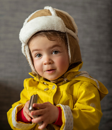 Cute little girl wrapped up with warm clothing, holding a phone in her hands. she is looking at camera with a cute face . Girl is dressed ina yellow jacket and a brown hat