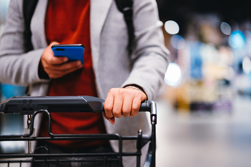 Anonymous man holding his smart phone and writing a text message while pushing a shopping cart in the supermarket.