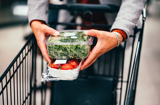 Unrecognizable man holding plastic boxes with vegetables and putting them into a shopping cart in the supermarket.