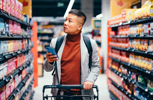 apuesto hombre asiático buscando comestibles de la lista en su teléfono móvil - retail people customer shopping fotografías e imágenes de stock