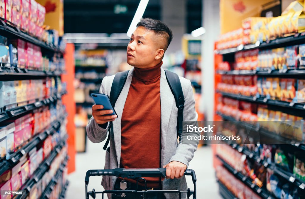 Handsome Asian Male Searching for Groceries From the List on His Mobile Phone Handsome Asian male walking down the product aisle in the supermarket, looking at shelves and searching for groceries from the list on his mobile phone he is holding in his hand. Retail Stock Photo