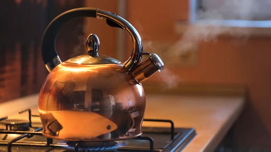 A metal silver teapot on a gas stove in the kitchen at home. Close up of steel kettle with boiling water, preparation of hot beverage.