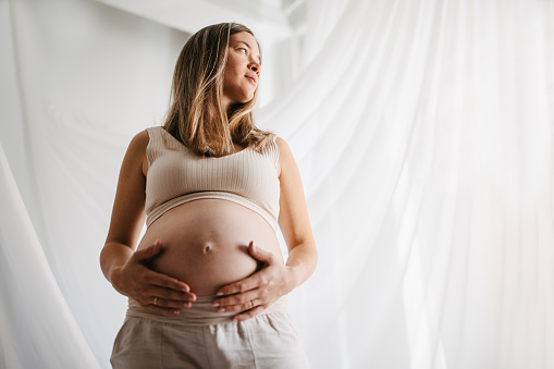 Asian beautiful and happy woman in pregnancy sitting beside the bed in bedroom. Satisfaction and relaxation of pregnant woman. 20-24 weeks belly of happy pregnant woman portrait with copyspace.