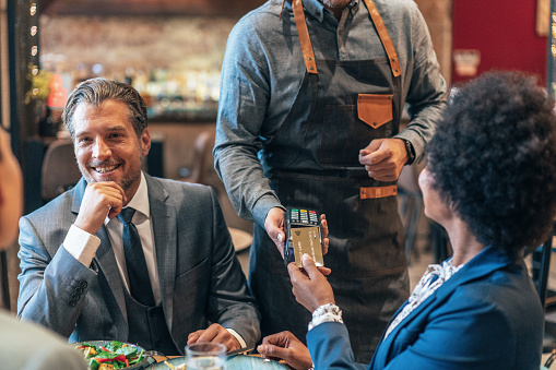 Beautiful African American woman paying for a lunch with business colleagues. A diverse group of businesspeople enjoying lunch at a restaurant, and an African-American woman paying with a credit card via contactless payment