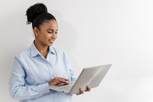 Studio portrait of young African American woman standing against a white wall and using a laptop
