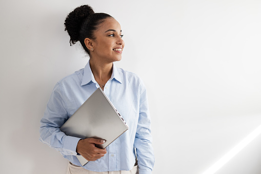 Young African American woman in blue shirt holding laptop. She is standing against white wall and looking away.