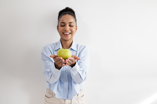 Smiling African American woman standing in front of the white wall and holding an apple. Healthy snack. Healthy diet.