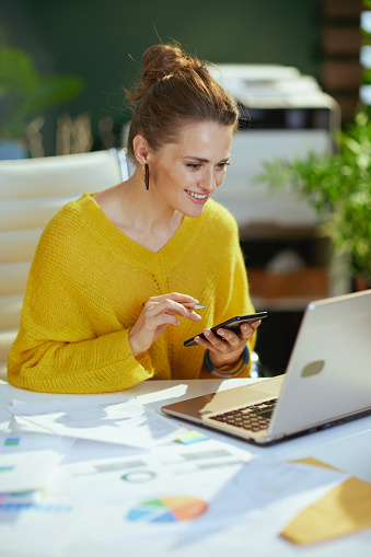 happy elegant small business owner woman in yellow sweater with laptop using smartphone in the modern green office.