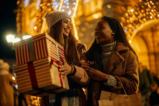 Two smiling multiracial female friends in Christmas shopping holding presents, shopping bags in decorated city