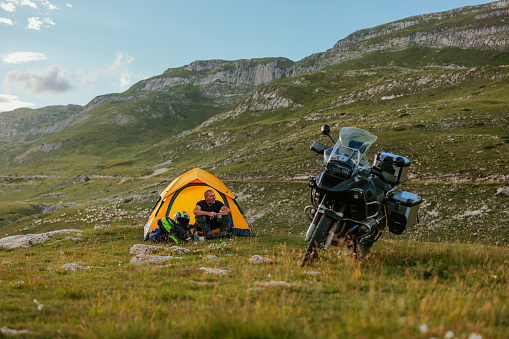 Biker is sitting in front of his tent in the mountains with his bike parked close by as the sun is rising. He is drinking coffee