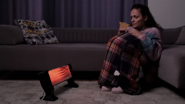 Person sitting on the floor heating their hands at home over a portable electric halogen heater in winter